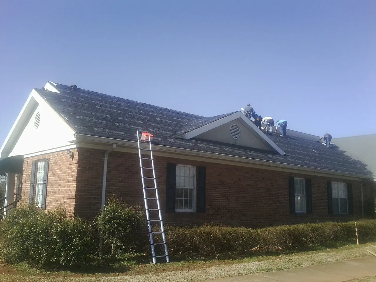 Crew tearing shingles off a brick ranch roof with a ladder set — Emergency Roof Repair work by our crew in Clarks Grove