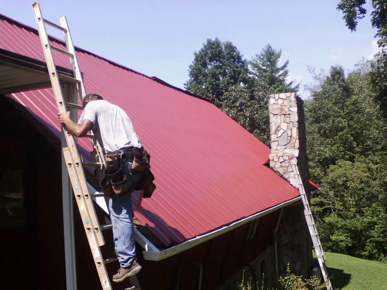 Roofer on a ladder beside a red metal roof and stone chimney — Soffit & Fascia Repair by Roofing Brothers in Clarks Grove
